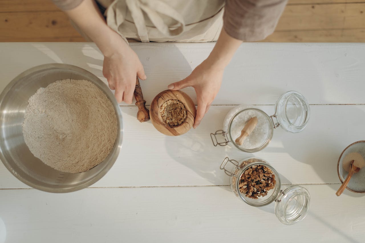Hands preparing baking ingredients with nuts and flour on a white table.